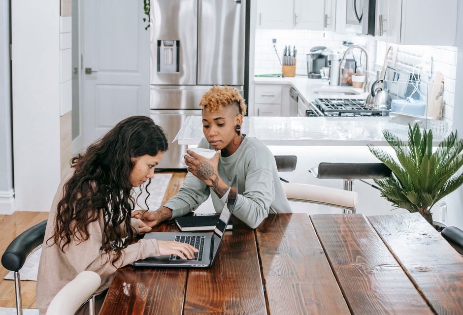A couple sits together at a sunlit kitchen table, looking at a laptop and documents, proactively and calmly managing their home finances together.